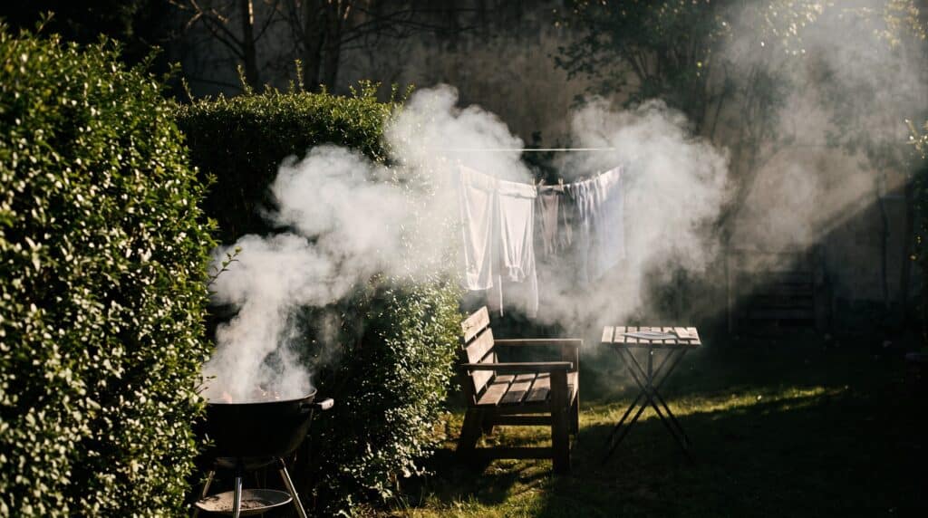 Dichter Rauch eines Holzkohlegrills zieht über eine Gartenhecke in den Nachbargarten mit Wäscheleine