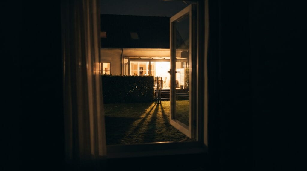 Blick aus einem dunklen Schlafzimmer durch das Fenster auf die hell erleuchtete Terrasse des Nachbarhauses bei Nacht
